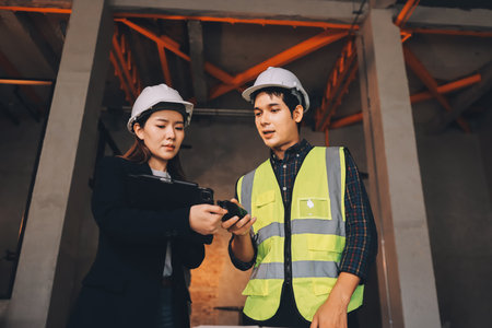 Construction manager and engineer dressed in orange work vests and hard helmets explore construction...