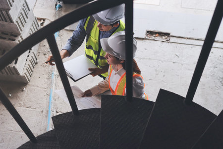 Construction manager and engineer dressed in orange work vests and hard helmets explore construction...
