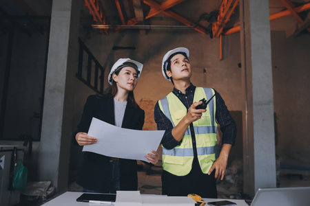 Construction manager and engineer dressed in orange work vests and hard helmets explore construction...