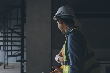 Construction manager and engineer dressed in orange work vests and hard helmets explore construction...
