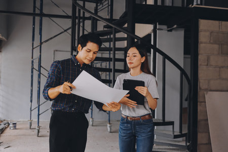 Construction manager and engineer dressed in orange work vests and hard helmets explore construction documentation on the building site near the steel framesの写真素材