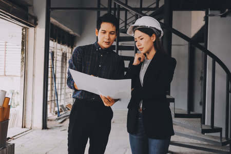 Construction manager and engineer dressed in orange work vests and hard helmets explore construction documentation on the building site near the steel framesの写真素材