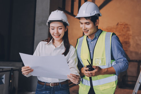 Construction manager and engineer dressed in orange work vests and hard helmets explore construction documentation on the building site near the steel framesの写真素材