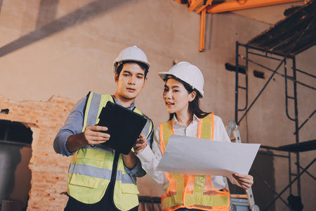 Construction manager and engineer dressed in orange work vests and hard helmets explore construction documentation on the building site near the steel framesの写真素材