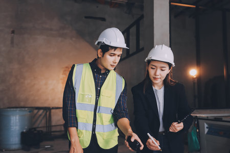 Construction manager and engineer dressed in orange work vests and hard helmets explore construction documentation on the building site near the steel framesの写真素材