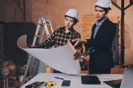 Construction manager and engineer dressed in orange work vests and hard helmets explore construction documentation on the building site near the steel framesの写真素材
