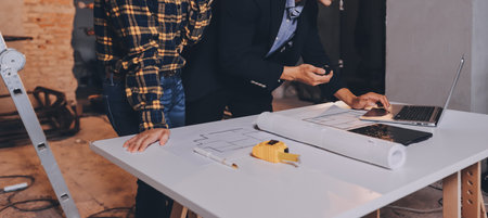 Construction manager and engineer dressed in orange work vests and hard helmets explore construction documentation on the building site near the steel framesの写真素材