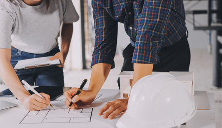Construction manager and engineer dressed in orange work vests and hard helmets explore construction documentation on the building site near the steel framesの写真素材