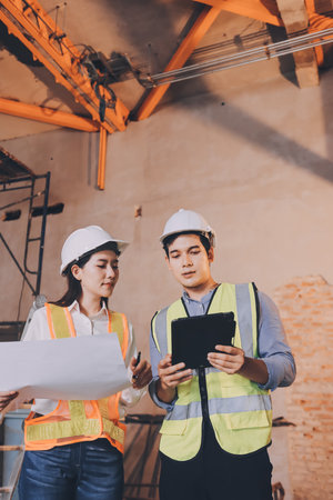 Construction manager and engineer dressed in orange work vests and hard helmets explore construction documentation on the building site near the steel framesの写真素材