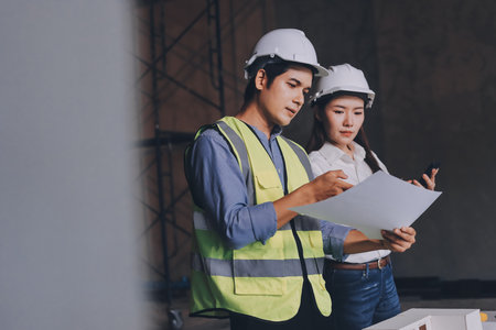 Construction manager and engineer dressed in orange work vests and hard helmets explore construction documentation on the building site near the steel framesの写真素材