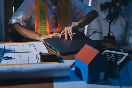 Closeup of an industrial engineer working on a laptop, with blueprints and a model house on the desk.の写真素材