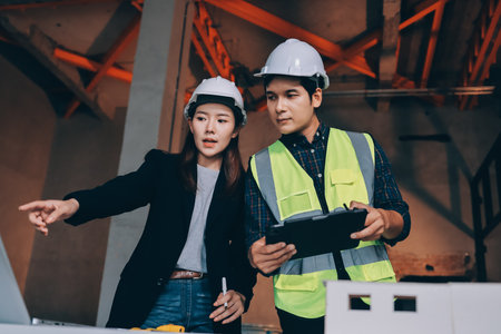 Construction manager and engineer dressed in orange work vests and hard helmets explore construction documentation on the building site near the steel framesの写真素材