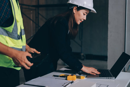 Construction manager and engineer dressed in orange work vests and hard helmets explore construction documentation on the building site near the steel framesの写真素材