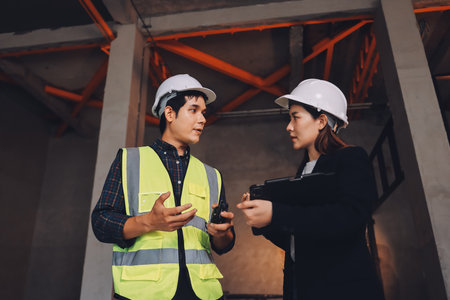Construction manager and engineer dressed in orange work vests and hard helmets explore construction...