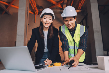 Construction manager and engineer dressed in orange work vests and hard helmets explore construction...