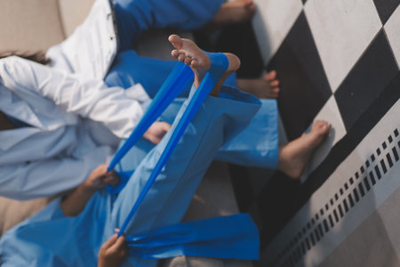 Confident physical therapist helps patient use resistance band stretching out his leg in clinic...