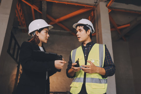 Construction manager and engineer dressed in orange work vests and hard helmets explore construction...