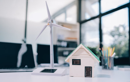 Close up of a wind turbine model and a house model on a desk in an office setting, with laptops and pencils in the background.の写真素材