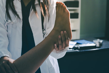 Doctor examining and supporting injured ankle of female patient in hospital room, providing medical assistance and treatment for sprain or fractureの写真素材