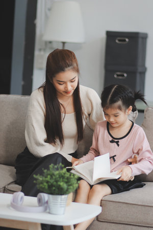A cheerful Asian mother helps her young daughter do homework and color a book at home, while the...