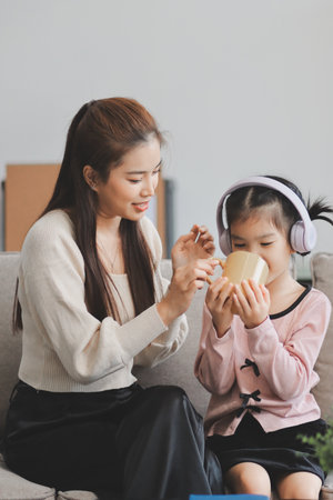 A cheerful Asian mother helps her young daughter do homework and color a book at home, while the father works in the background. A cozy, bright, and modern family lifestyle.の写真素材