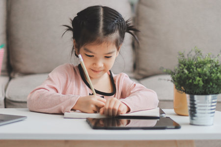 A cheerful Asian mother helps her young daughter do homework and color a book at home, while the...