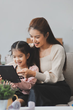 A cheerful Asian mother helps her young daughter do homework and color a book at home, while the father works on a laptop in the background. A cozy, bright, and modern family lifestyle.の写真素材