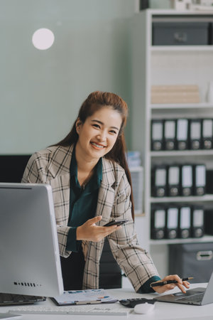 Determined asian businesswoman works late in a modern office, confidently analyzing data on her laptop, embodying success and professionalism with a smileの写真素材