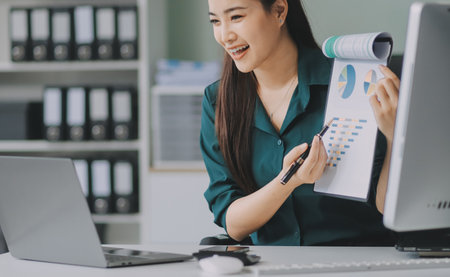 Determined asian businesswoman works late in a modern office, confidently analyzing data on her laptop, embodying success and professionalism with a smileの写真素材
