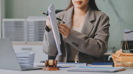Confident Lawyer Working at Desk: A young Asian female lawyer confidently works on her laptop, surrounded by legal symbols. She exudes professionalism and competence in her modern office setting.の写真素材