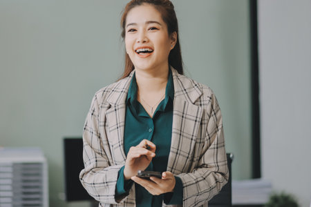 Determined asian businesswoman works late in a modern office, confidently analyzing data on her laptop, embodying success and professionalism with a smileの写真素材
