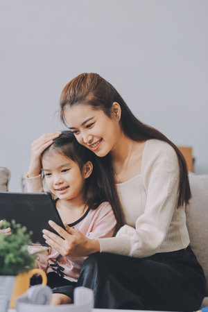 A cheerful Asian mother helps her young daughter do homework and color a book at home, while the father works in the background. A cozy, bright, and modern family lifestyle.の写真素材