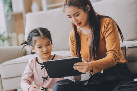 A cheerful Asian mother helps her young daughter do homework and color a book at home, while the...