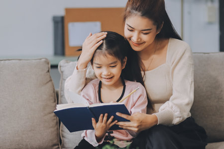 A cheerful Asian mother helps her young daughter do homework and color a book at home, while the father works on a laptop in the background. A cozy, bright, and modern family lifestyle.の写真素材