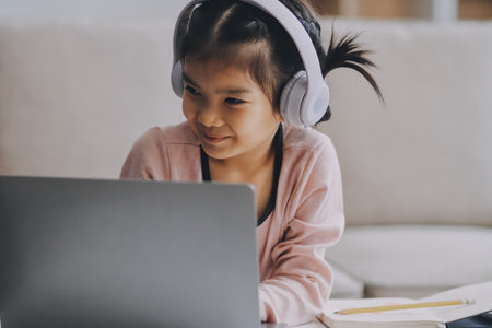 A cheerful Asian mother helps her young daughter do homework and color a book at home, while the father works on a laptop in the background. A cozy, bright, and modern family lifestyle.の写真素材