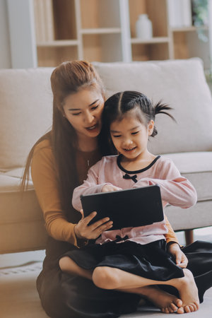 A cheerful Asian mother helps her young daughter do homework and color a book at home, while the...
