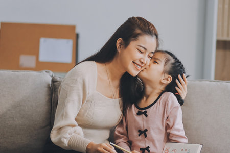A cheerful Asian mother helps her young daughter do homework and color a book at home. A cozy, bright, and modern family lifestyle.の写真素材