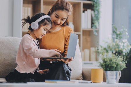 A cheerful Asian mother helps her young daughter do homework and color a book at home, while the father works on a laptop in the background. A cozy, bright, and modern family lifestyle.の写真素材