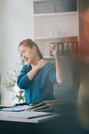 Asian businesswoman experiencing neck and shoulder pain, stretching during long hours working on...
