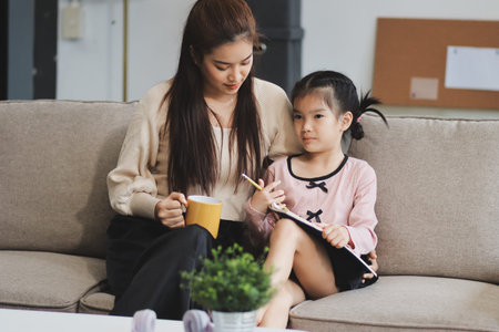 A cheerful Asian mother helps her young daughter do homework and color a book at home, while the father works on a laptop in the background. A cozy, bright, and modern family lifestyle.の写真素材