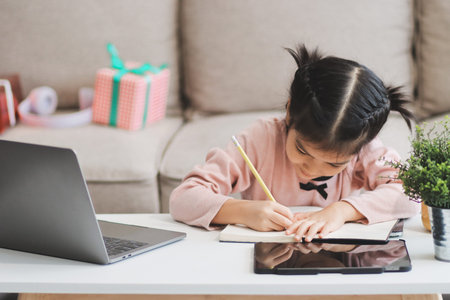 A cheerful Asian mother helps her young daughter do homework and color a book at home, while the...