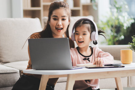 A cheerful Asian mother helps her young daughter do homework and color a book at home, while the...
