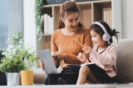 A cheerful Asian mother helps her young daughter do homework and color a book at home, while the...