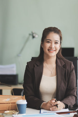 Confident Lawyer Working at Desk: A young Asian female lawyer confidently works on her laptop, surrounded by legal symbols. She exudes professionalism and competence in her modern office setting.の写真素材