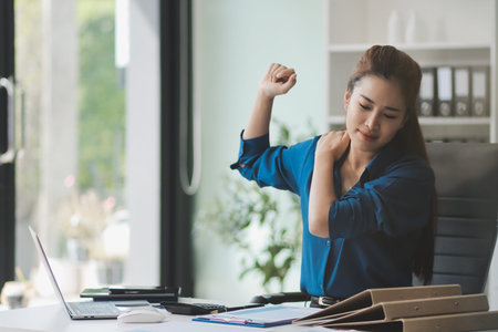 Asian businesswoman experiencing neck and shoulder pain, stretching during long hours working on...