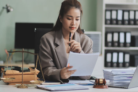 Confident Lawyer Working at Desk: A young Asian female lawyer confidently works on her laptop, surrounded by legal symbols. She exudes professionalism and competence in her modern office setting.の写真素材