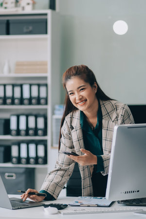 Determined asian businesswoman works late in a modern office, confidently analyzing data on her laptop, embodying success and professionalism with a smileの写真素材