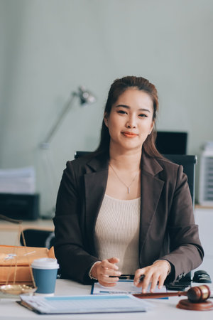 Confident Lawyer Working at Desk: A young Asian female lawyer confidently works on her laptop, surrounded by legal symbols. She exudes professionalism and competence in her modern office setting.の写真素材