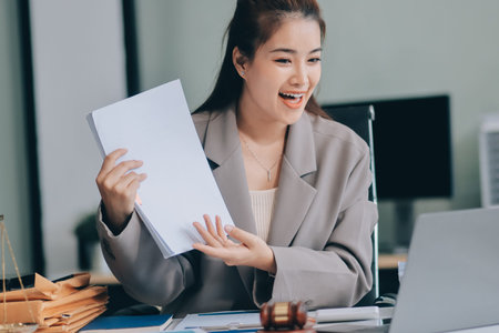 Confident Lawyer Working at Desk: A young Asian female lawyer confidently works on her laptop, surrounded by legal symbols. She exudes professionalism and competence in her modern office setting.の写真素材