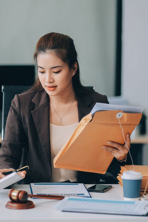 Confident Lawyer Working at Desk: A young Asian female lawyer confidently works on her laptop, surrounded by legal symbols. She exudes professionalism and competence in her modern office setting.の写真素材